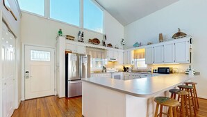 A modern kitchen with white cabinetry, a large island with stools, stainless steel appliances, and decorative items on the upper shelves. - Spacious oceanfront home w/ stunning views, large deck - private access to beach (Folly Beach)