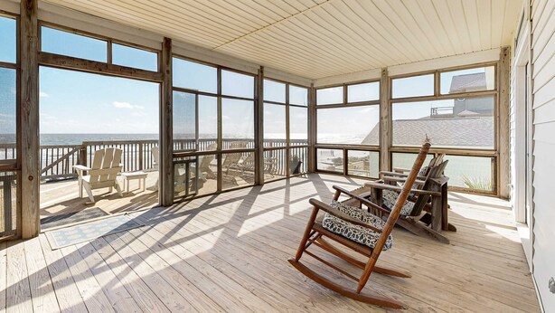 A covered deck with wooden flooring, large windows, and outdoor furniture including chairs and a rocking chair. - Spacious oceanfront home w/ stunning views, large deck - private access to beach (Folly Beach)