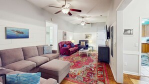 A living room with a grey sectional sofa, a red armchair, and a patterned rug. There are ceiling fans and wall-mounted artwork.