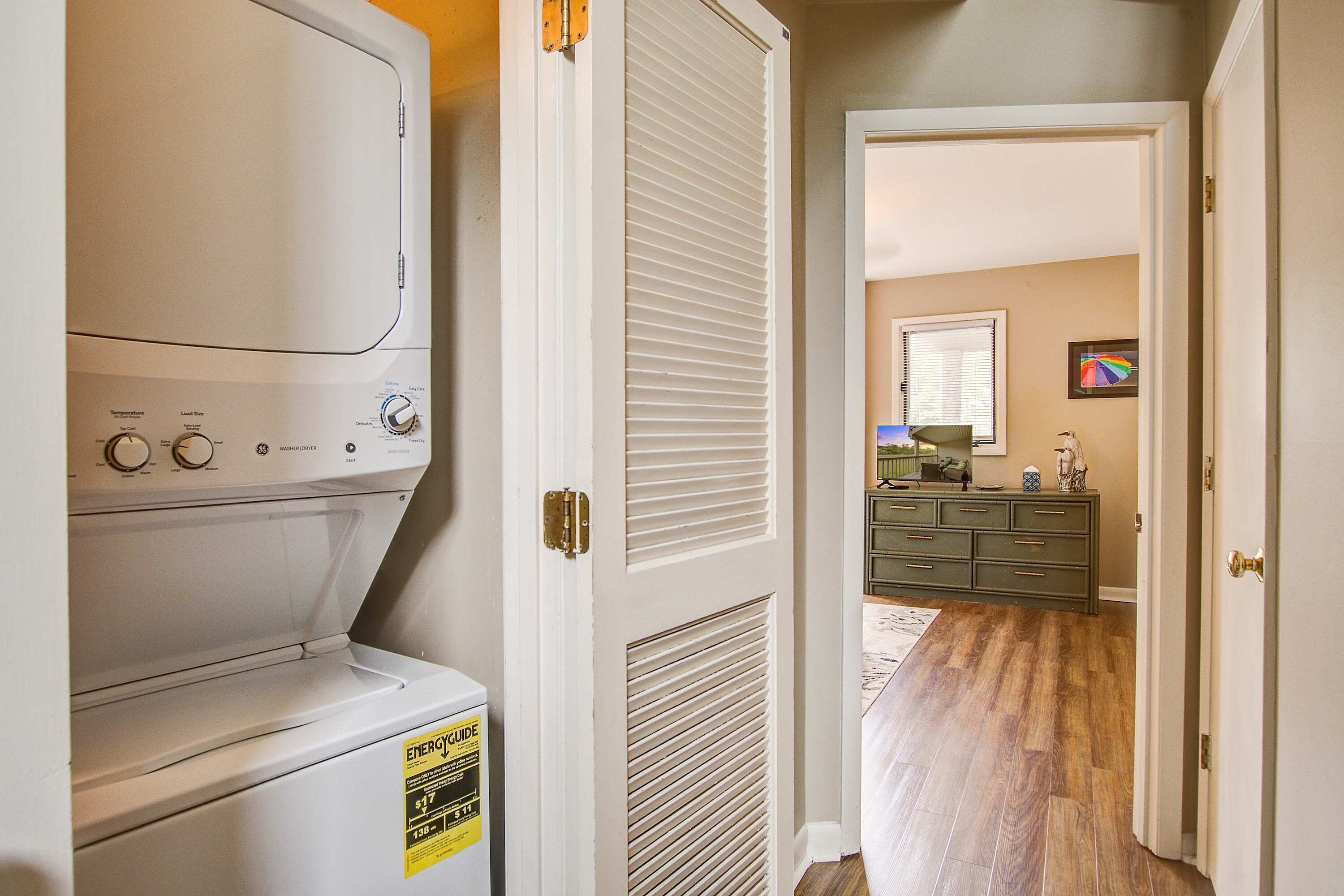 A laundry room with a front-loading washer and dryer, a door leading to a hallway with a dresser and a mirror.
