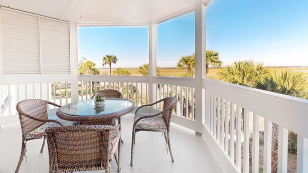 A covered balcony with wicker furniture and a glass table, overlooking a beach with palm trees. - Marsh-view condo with on-site pool, tennis, & private balcony (Folly Beach)