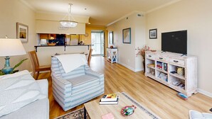 A living room with a sofa, a patterned armchair, a coffee table with books, a TV stand, and a kitchen in the background. - Marsh-view condo with on-site pool, tennis, & private balcony (Folly Beach)
