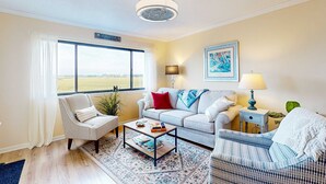A living room with a sofa, armchair, coffee table, and a large window with a view of the countryside. - Marsh-view condo with on-site pool, tennis, & private balcony (Folly Beach)