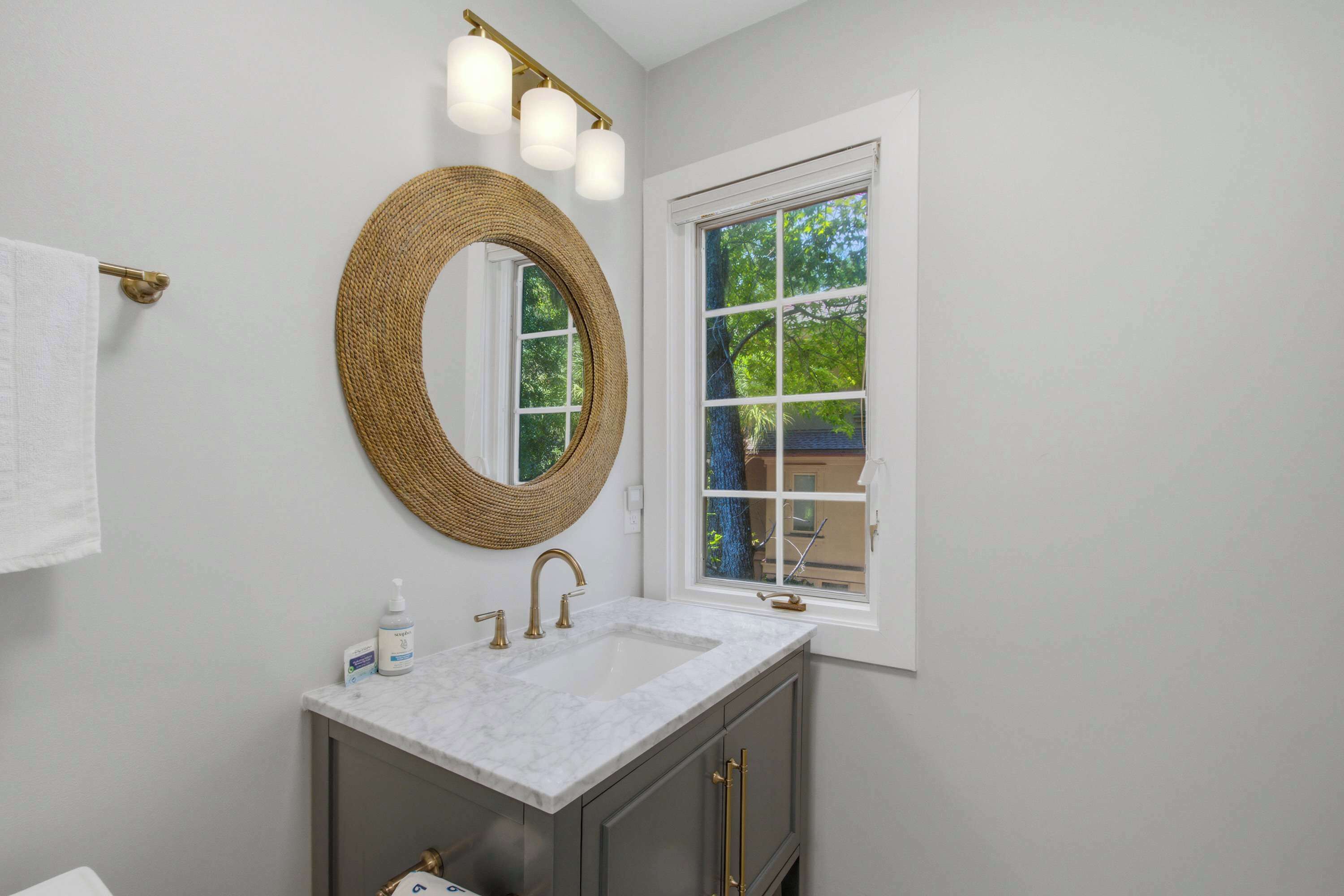 A bathroom with a round mirror, a white sink, and a window with a view of trees.
