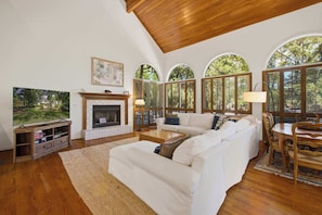 A spacious living room with a fireplace, white sofas, a television, and large windows with views of trees.