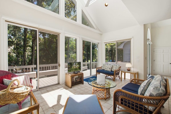 A sunlit room with a large sliding glass door leading to a deck, wicker furniture, and a television on a wooden stand.