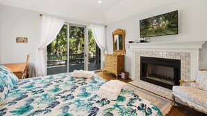 A bedroom with a bed, a fireplace, a television, and a balcony with a view of trees.