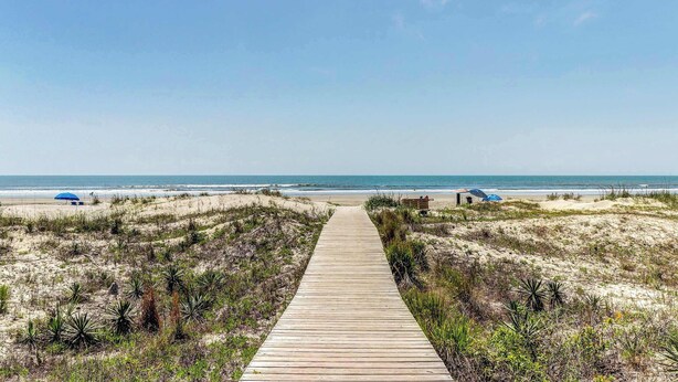 Een houten looppad dat naar een strand leidt, met duinen en schaarse begroeiing.