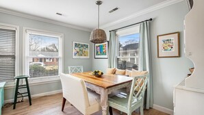 A dining area with a wooden table, white upholstered chairs, a wicker pendant light, and framed artwork on the walls. - Darling, renovated home w/ beautiful backyard, W/D & WiFi - near beaches (Mt. Pleasant)