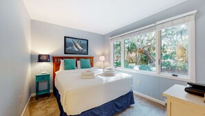 Een slaapkamer met een groot bed, een nachtkastje, een lamp, een lijst met een afbeelding en een raam met uitzicht op groen.