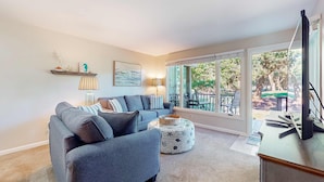 Een moderne woonkamer met een grijze hoekbank, een rond poef met een zeesterrenmotief, en een groot raam met uitzicht op tuinmeubilair.