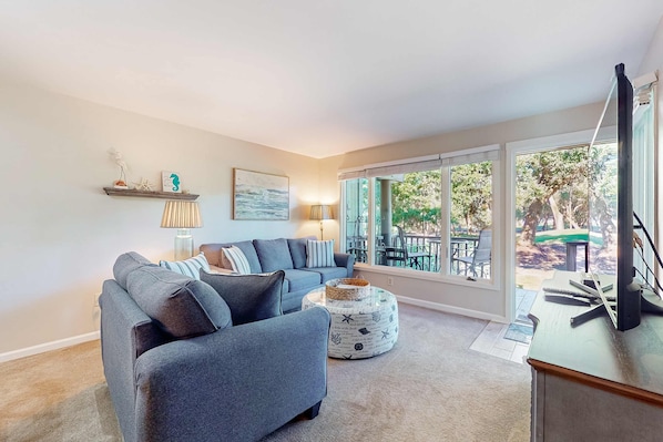 A modern living room with a grey sectional sofa, a round ottoman with a starfish pattern, and a large window offering a view of outdoor furniture. - Five minute walk to beautiful beaches, golf course views, pet friendly (Kiawah Island)