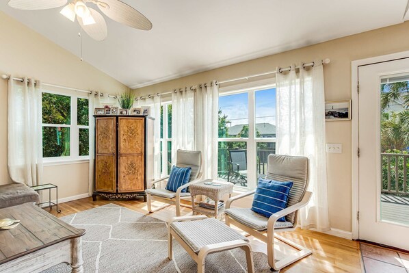 A sunlit room with a wooden cabinet, two chairs with cushions, a small table, and a large window with curtains. - Relaxing oceanside home w/ multiple decks, outdoor shower- steps to beach access (Folly Beach)