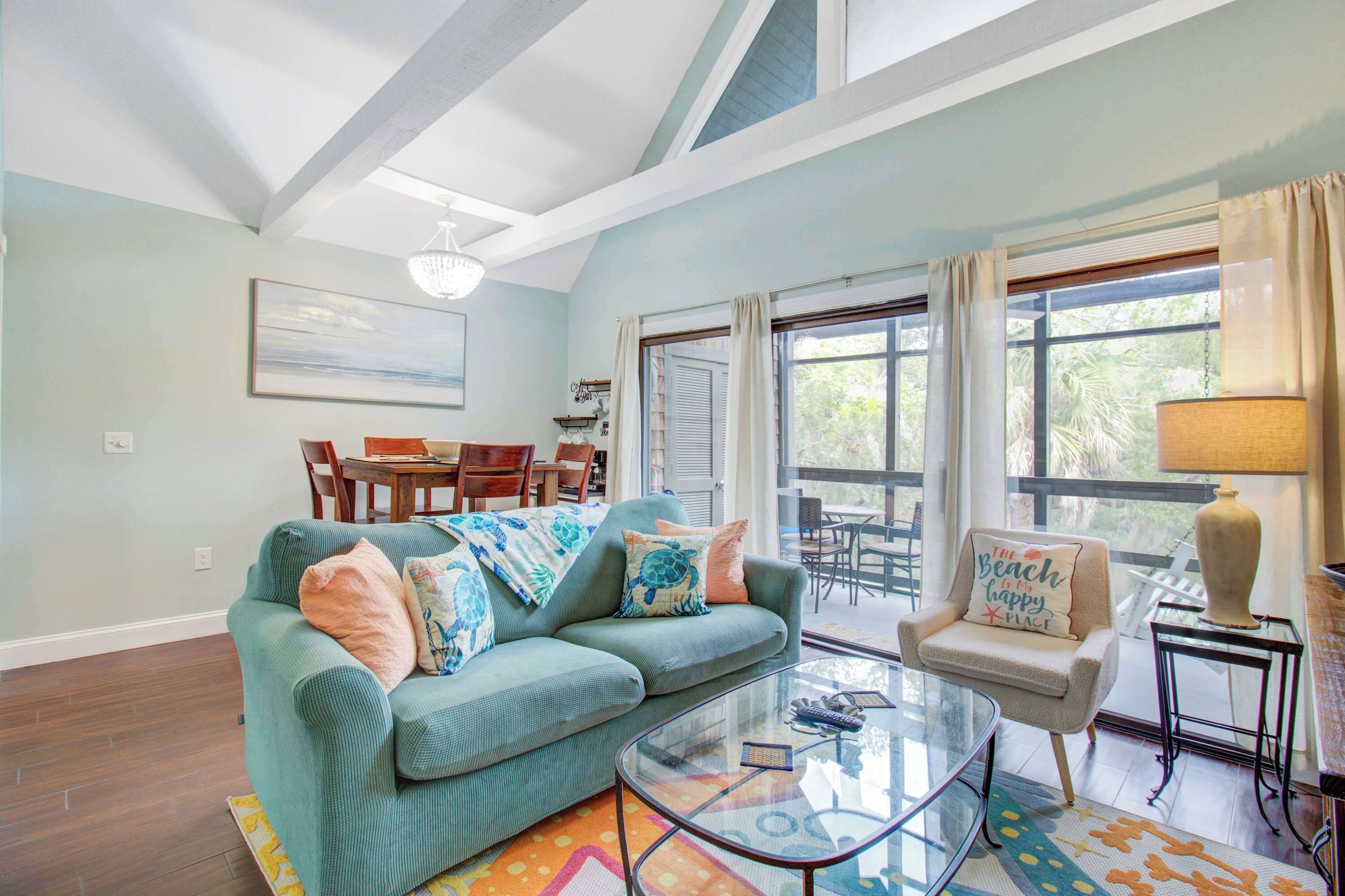 A living room with a teal sofa, a glass coffee table, and a chair with a beach-themed pillow.