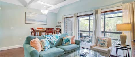 A living room with a teal sofa, a glass coffee table, and a chair with a beach-themed pillow.