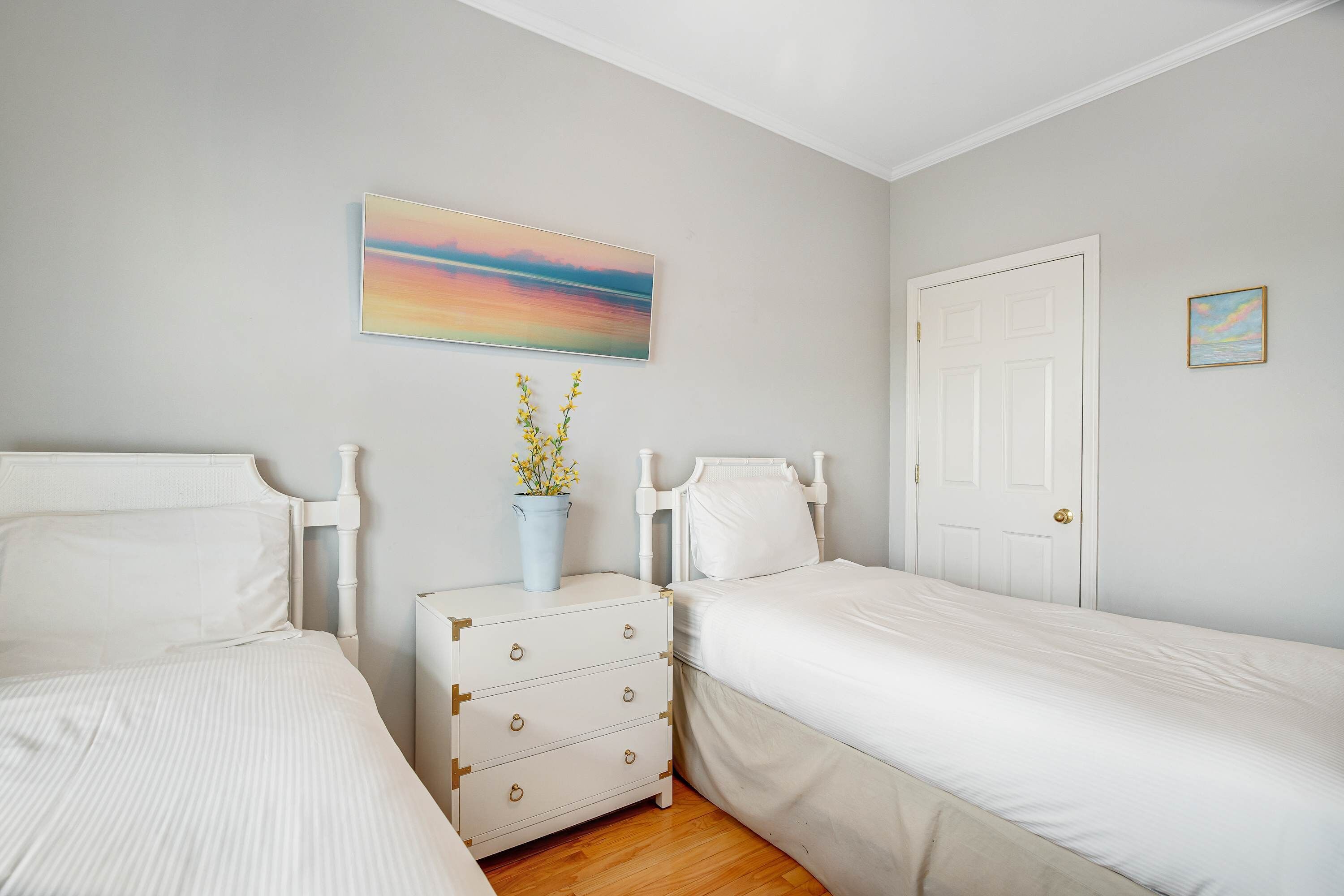 Een slaapkamer met twee eenpersoonsbedden, een nachtkastje met lades, een witte deur en een schilderij aan de muur.