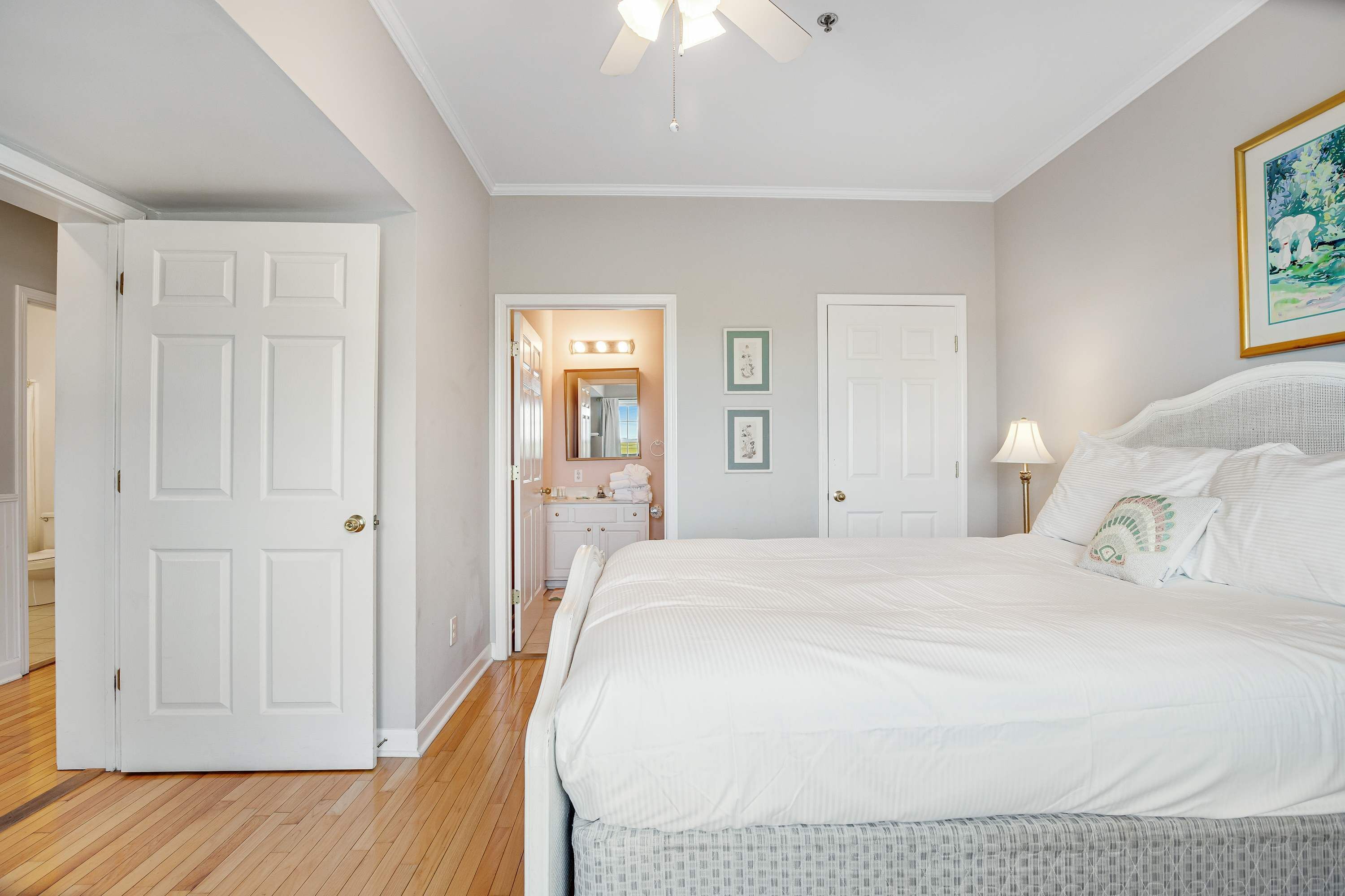 Une chambre à coucher avec un grand lit, un ventilateur de plafond et une salle de bain visible à travers une porte ouverte.