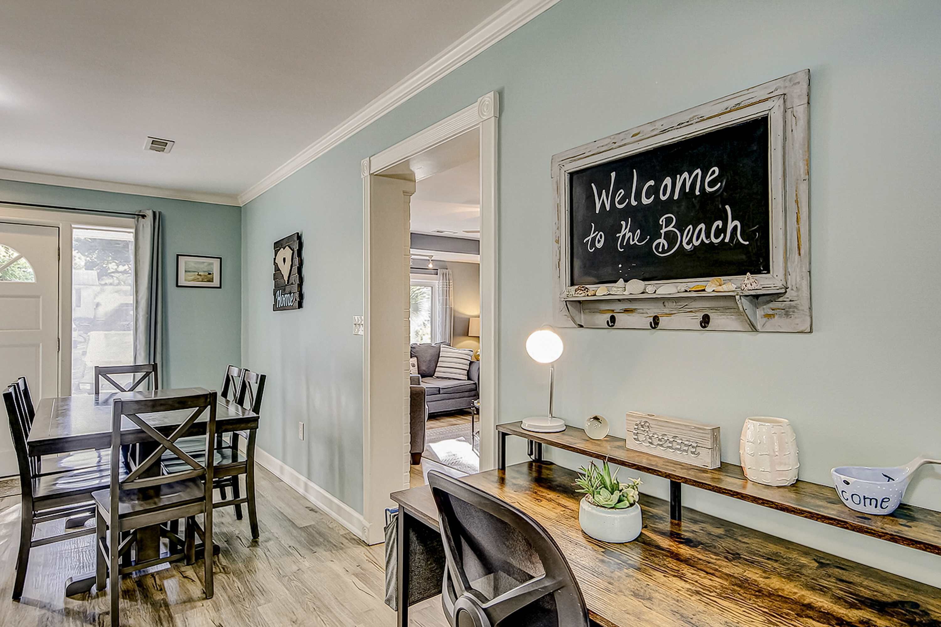 A dining area with a wooden table and chairs, a chalkboard sign saying "Welcome to the Beach", and a view into another room.