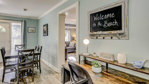Un coin repas avec une table et des chaises en bois, un panneau noir sur lequel est écrit « Bienvenue à la plage », et une vue sur une autre pièce.