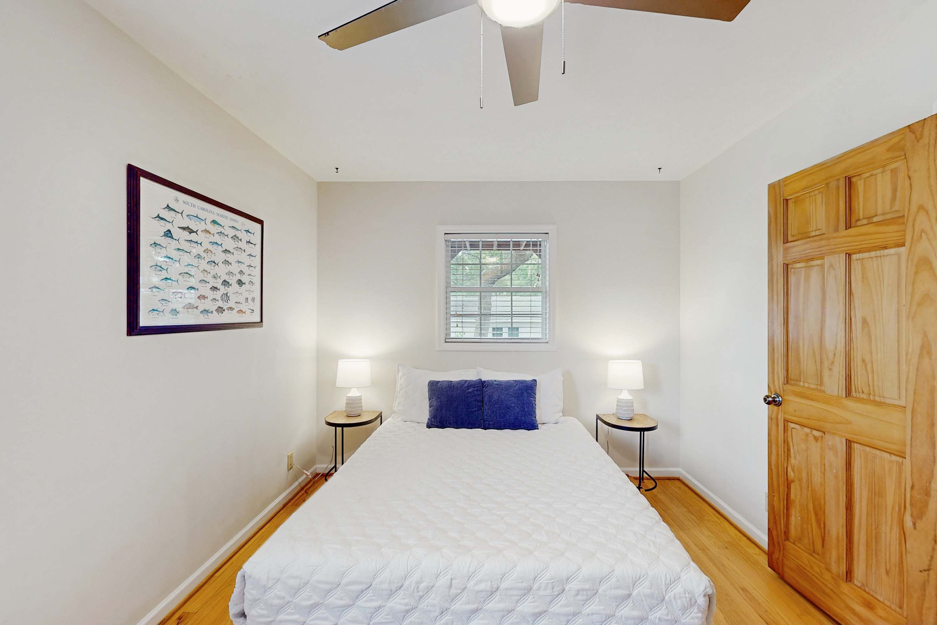 A bedroom with a bed, two bedside tables, a window, a ceiling fan, and a wooden door.