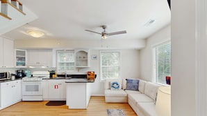 A modern kitchen with white cabinets, a built-in oven, and a central island. There is a white sofa with patterned cushions and a window with blinds. - Amazing Beach Home (Folly Beach)