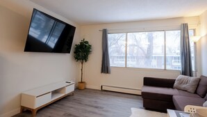 Una sala de estar moderna con un televisor de pantalla plana montado en la pared, un sofá marrón, un banco blanco y un amplio ventanal con cortinas.