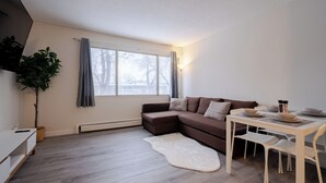 Una sala de estar moderna con un sofá seccional marrón, una mesita blanca con platos y una lámpara, un tapete blanco y una planta en maceta.