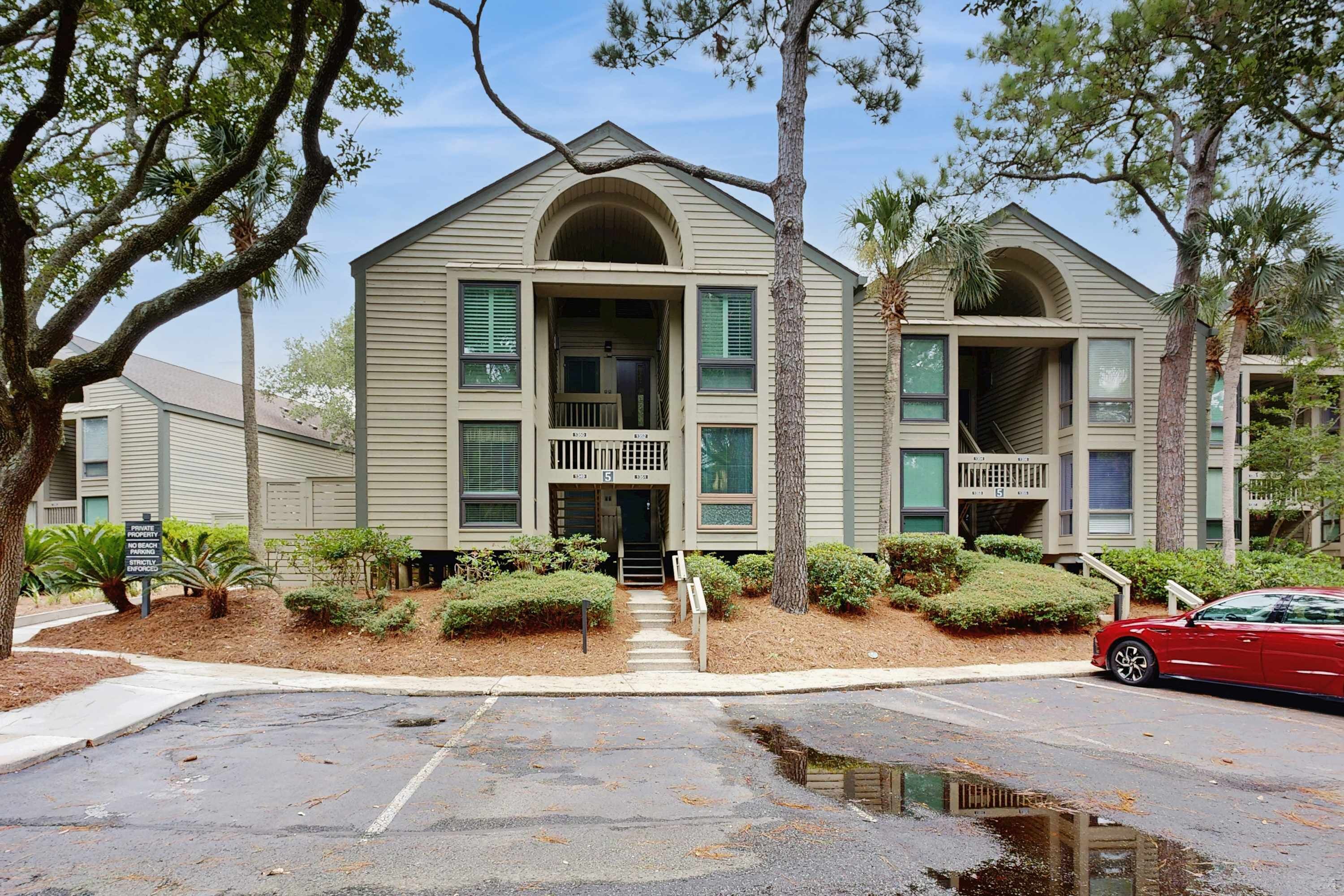 A two-story building with a central entrance, a small porch, and a parking lot with a red car.