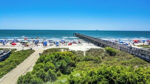 A beach with a wooden pier extending into the ocean, surrounded by greenery and beach umbrellas. - Ocean front condo villa near restaurants & state park with beach access, pool & pier (Isle of Palms)