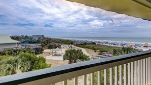 A beach view from a balcony with a railing, palm trees, and beach umbrellas. - Ocean front condo villa near restaurants & state park with beach access, pool & pier (Isle of Palms)