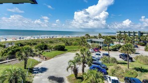 A parking lot with several cars, a building with a green roof, and a beach with umbrellas in the background. - Ocean front condo villa near restaurants & state park with beach access, pool & pier (Isle of Palms)