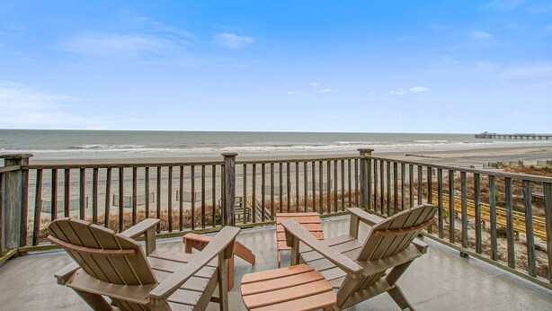 Een balkon met twee Adirondack-stoelen en een tafeltje, met uitzicht op een strand en pier.