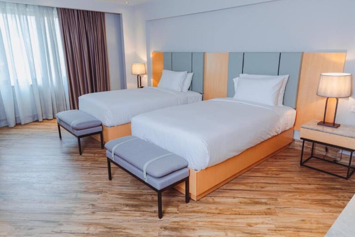 A hotel room with two beds, a bench, and a nightstand with lamps.