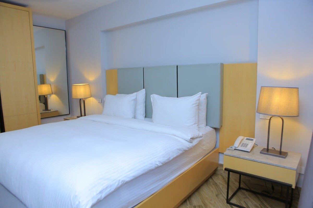 A neatly made bed with white linens, a bedside table with a lamp and a phone, and a mirror on the wall.