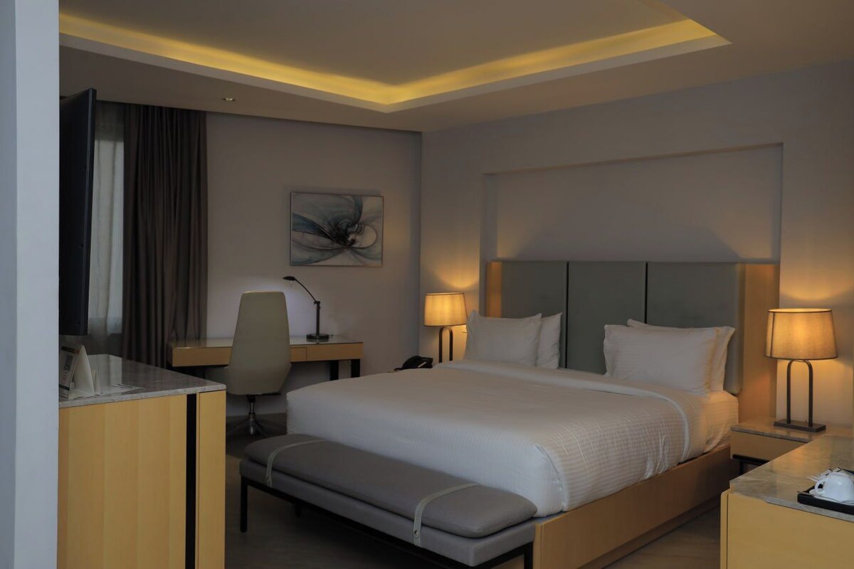 Een hotelkamer met een groot bed, bedlampen, een bureau met een stoel, een bankje en een schilderij aan de muur.