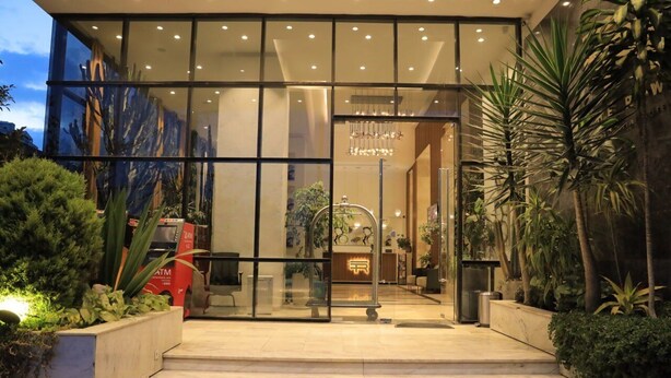 Entrada moderna de hotel con puertas de vidrio, una escalera que sube y plantas de interior.