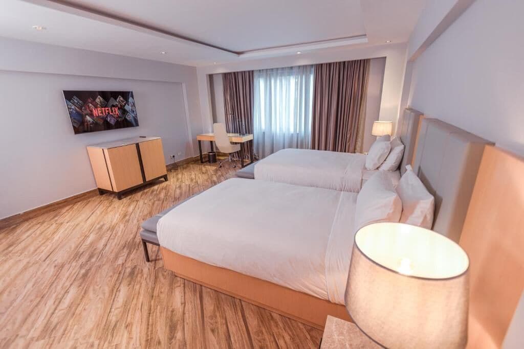A modern hotel room with a large bed, a desk, a chair, a TV mounted on the wall, and a window with curtains.