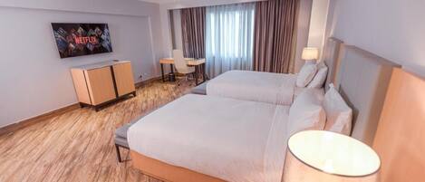 Una habitación de hotel moderna con una cama grande, un escritorio, una silla, un televisor montado en la pared y un ventanal con cortinas.