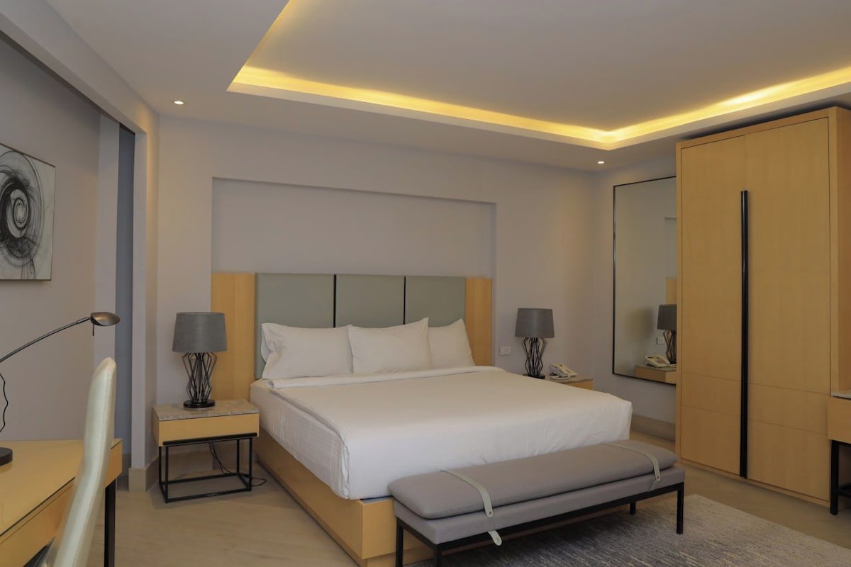 A modern hotel room with a large bed, bedside tables with lamps, a bench, a wardrobe, and a mirror.