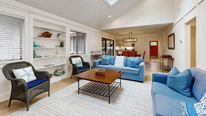 Ein geräumiges Wohnzimmer mit hoher Decke, einem Oberlicht, einem blauen Sofa, einem Korbstuhl und einem Holztisch.