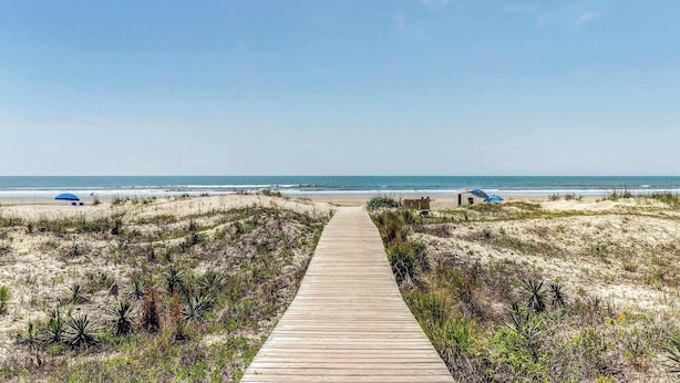 Ein hölzerner Steg führt zu einem Strand mit Dünen und spärlicher Vegetation.