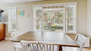 Un coin repas avec une table en bois et des chaises blanches, de grandes fenêtres équipées de stores, une décoration en forme d’étoile de mer et un petit tableau représentant un poisson accroché au mur.