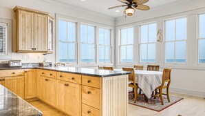 A kitchen with wooden cabinets, a granite countertop, and a dining area with a table and chairs. - Gorgeous Beachfront Home w/ Oceanfront Views, Deck, Beach Boardwalk & WiFi (Folly Beach)