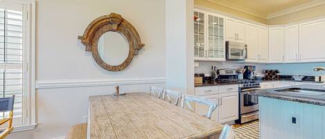 Un coin repas avec une table et des chaises en bois, un miroir rond accroché au mur, et une cuisine avec des meubles et des appareils électroménagers blancs.