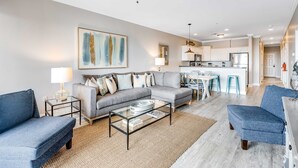 Una sala moderna con un sofá gris, sillones azules, una mesa de centro de vidrio y una cocina al fondo.