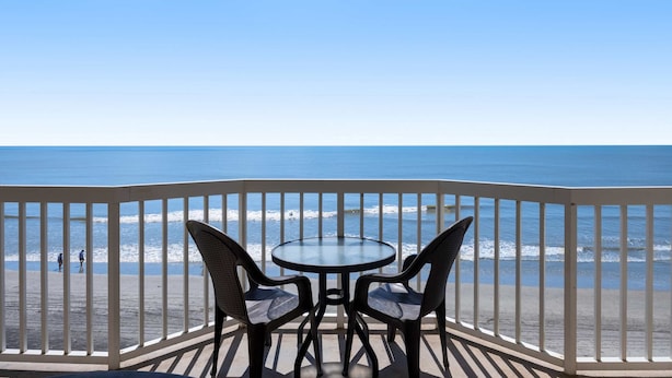 Een balkon met een glazen tafel en twee stoelen, met uitzicht op een strand waar mensen in de verte lopen.