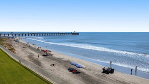 Een strand met een pier die het water in loopt, parasols en mensen op het strand.