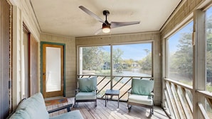 A covered porch with two chairs and a table, large windows, and a ceiling fan.