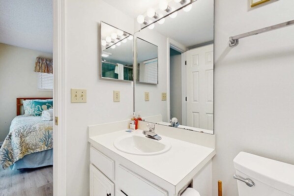A bathroom with a white sink, a large mirror, and a toilet. There is a bed with a blue and white patterned bedspread in the adjacent room. - First floor villa w/ lagoon views from the screened porch - close to Atlantic (Kiawah Island)