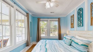 Une chambre avec un grand lit, un ventilateur de plafond et une vue sur les palmiers par la fenêtre.